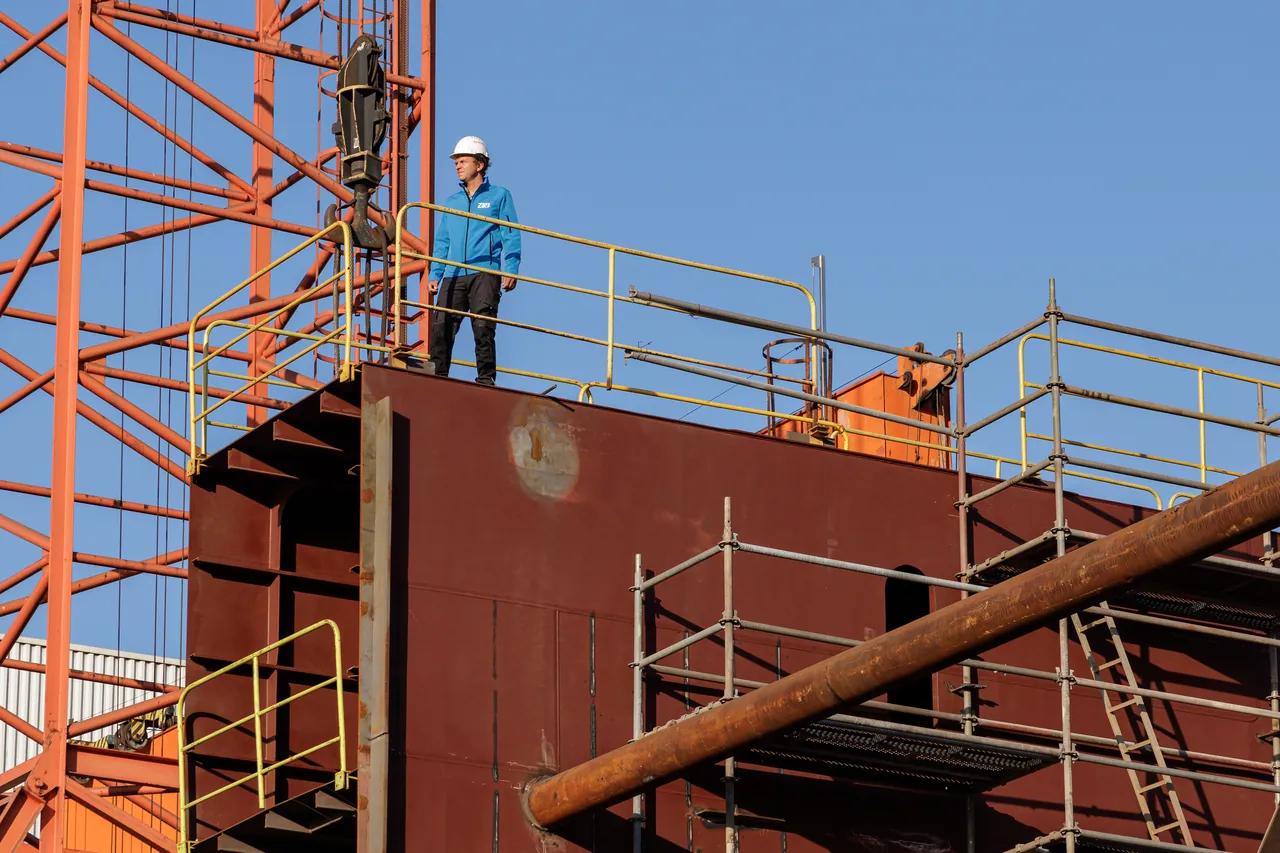 Un ouvrier debout sur un bloc de coque en acier, devant une grue du chantier naval