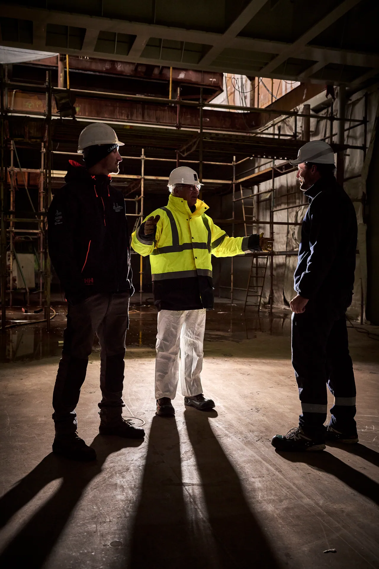 Trois hommes casqués en discussion à contre-jour dans la cale d’un navire en chantier
