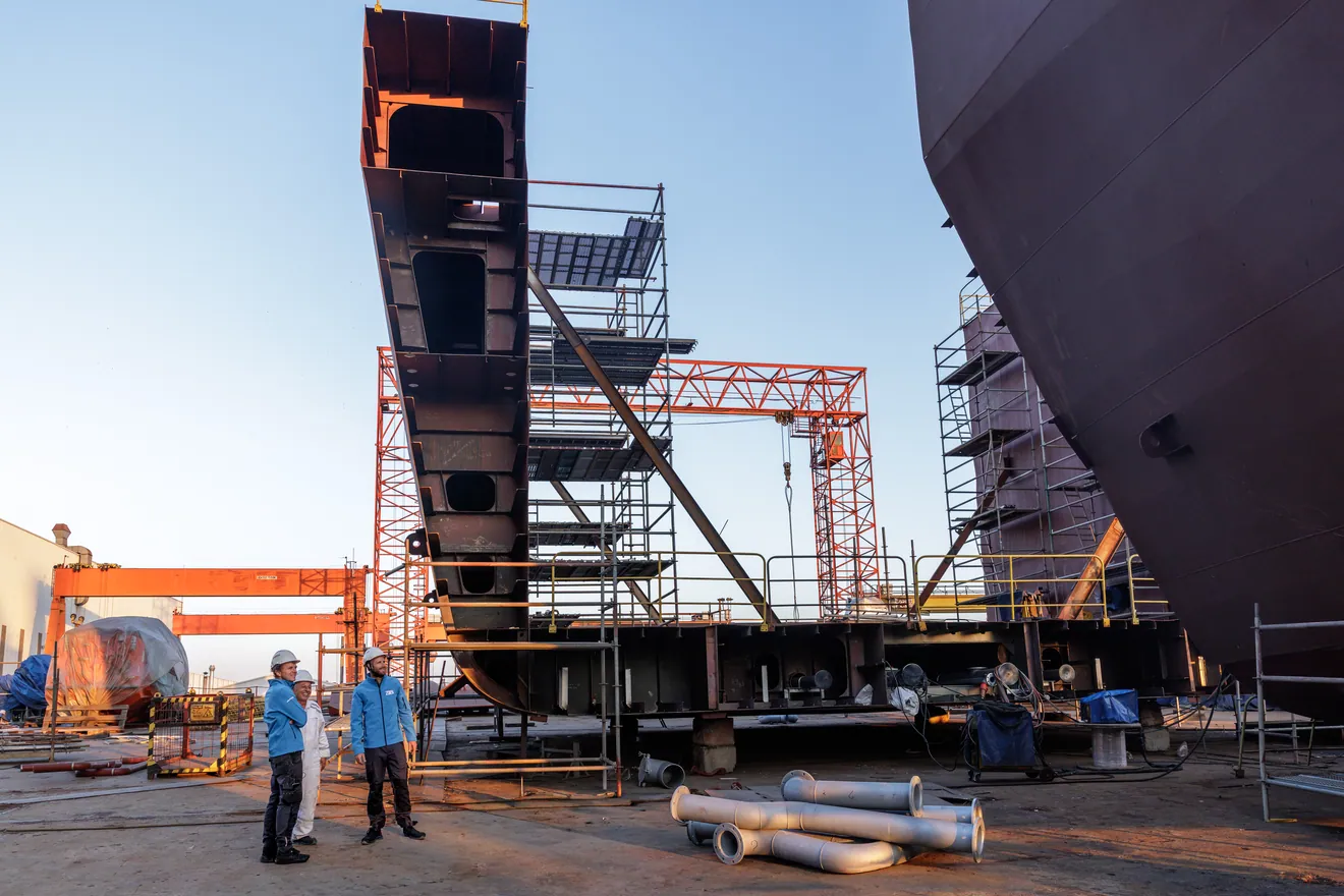 Sur le chantier du navire Windcoop, un bloc de coque levé par une grue, deux personnes au premier plan